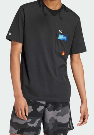 Person iført en sort Adidas T-shirt med tegneserieøjne og en flamme på lommen, kombineret med sorte og grå camouflage-shorts.