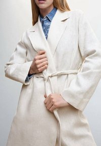 Cremefarvet linned coate med bindebælte, lange ærmer, klippet krave og subtil tekstur. Lagd over en denimskjorte, hvilket viser et afslappet look.