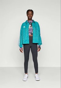 Jachetă teal cu un model texturat, colanți negri și un top scurt multicolor. Jacheta are dungi albe și logo-ul Adidas.