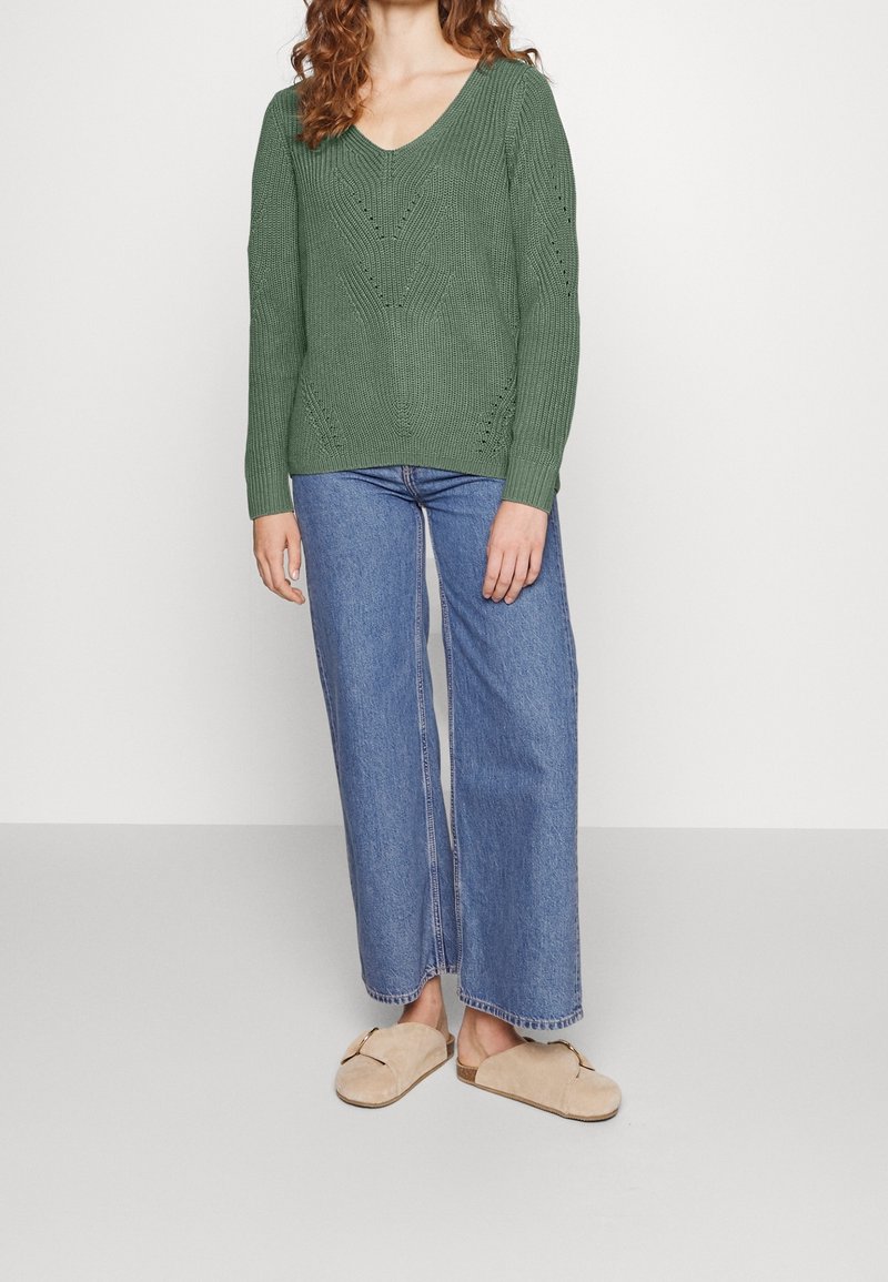 Femme portant un pull en maille vert à col en V, un jean large bleu et des chaussures beige à enfiler, debout devant un fond blanc uni.