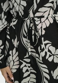 Musta kankainen mekko, jossa on suuret valkoiset lehtikuviot, sidottu vyötärö ja pitkät hihat, joissa on kerättyjä cuffeja. Teksturoitu pellavasekoitemateriaali.