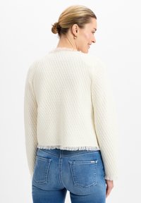 Cardigan bianco lavorato a maglia con un motivo testurizzato, lunghezza cropped e dettaglio a frange all'orlo. Indossato sopra jeans in denim blu.