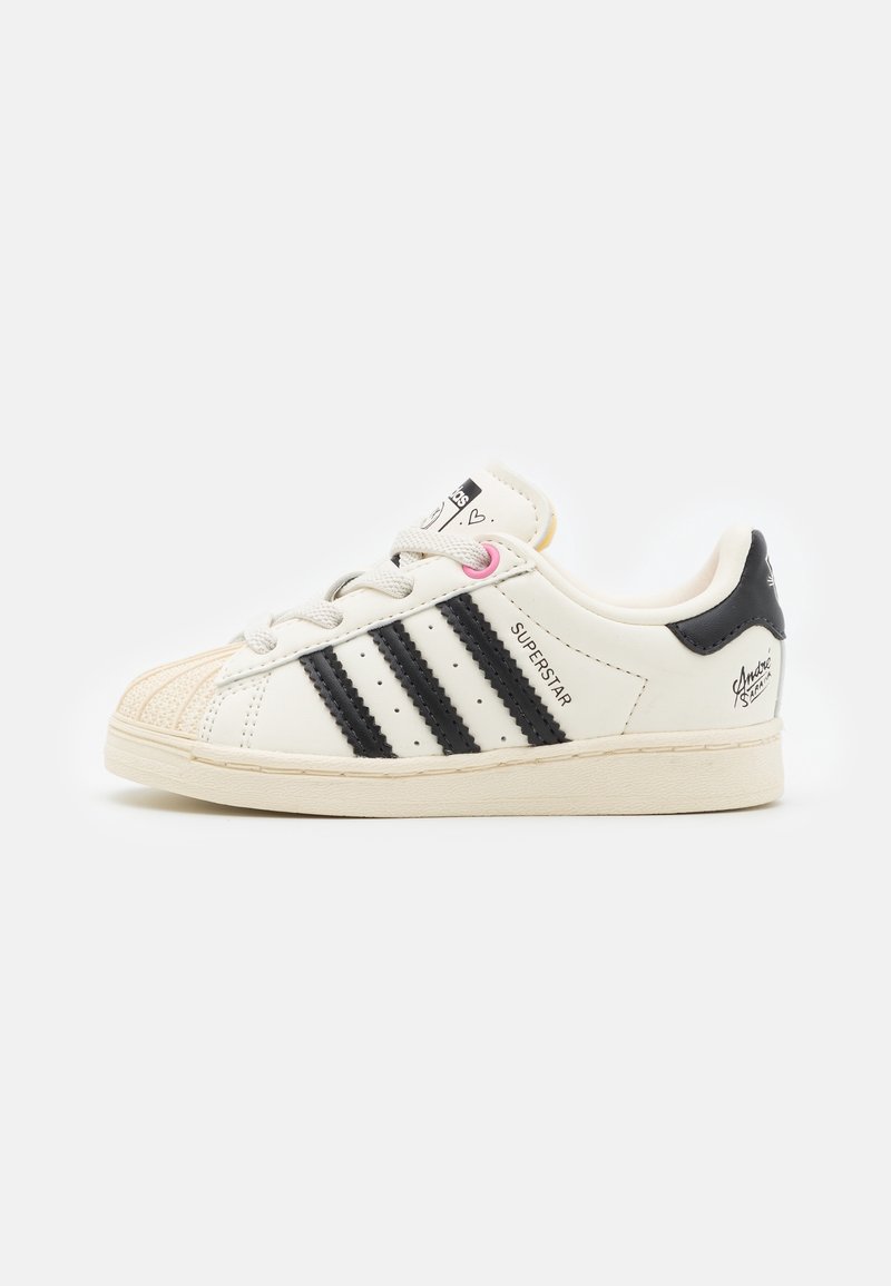 White leather sneaker with black stripes and accents, rubber sole, textured toe cap, and pink lace loop. "SUPERSTAR" logo on side.