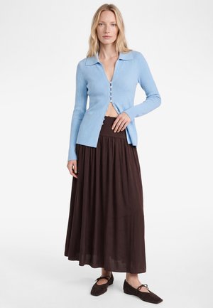 Frau trägt eine hellblaue, geknöpfte Strickjacke, einen langen dunkelbraunen Rock und dunkelbraune flache Schuhe, steht vor einem schlichten weißen Hintergrund.