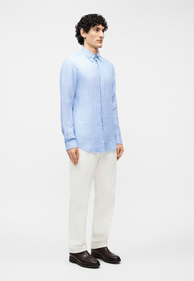 Άνδρας που στέκεται, φοράει ανοιχτό μπλε ριγέ πουκάμισο με κουμπιά, άσπρο παντελόνι και σκούρα καφέ παπούτσια, μπροστά σε απλό λευκό φόντο.