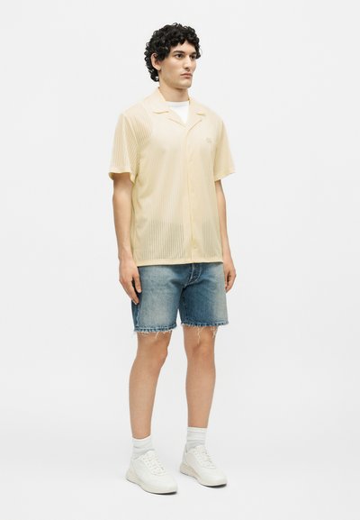 Νέος άνδρας στέκεται, φορώντας ένα ανοιχτό μπεζ κοντομάνικο πουκάμισο με κουμπιά, τζιν σορτσάκια, λευκές κάλτσες και λευκά αθλητικά παπούτσια.