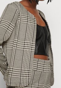 Blazer à carreaux noir et crème, de coupe courte avec ouverture devant. Porté sur un top en cuir noir. Tissu texturé avec des lignes nettes.