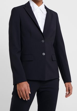 Kvinde iført en skræddersyet sort blazer med to knapper og klaplommer over en hvid skjorte med krave mod en ensfarvet baggrund.
