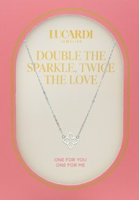 Zilveren ketting met een bloemvormige hanger, tentoongesteld op een roze kaart. Tekst: "DUBBEL DE GLANS, TWICE DE LIEFDE."