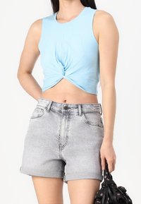 Světle modrý crop top s designem s překřížením vpředu, kombinovaný s šedými šortkami s vysokým pasem a ohrnutým lemem. Modelka drží černou kabelku.