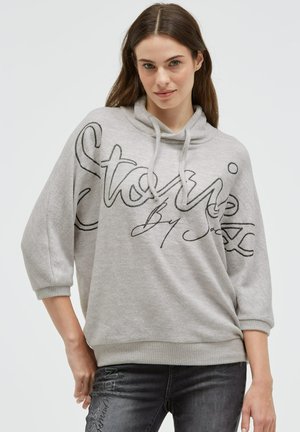 Junge Frau mit langen braunen Haaren, die einen hellgrauen Hoodie mit schwarzer Schrift und dunkelgraue Jeans vor einem schlichten weißen Hintergrund trägt.