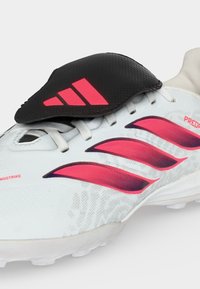 Λευκό ποδοσφαιρικό παπούτσι Adidas με κόκκινες ρίγες, μαύρο αναδιπλούμενο κάλυμμα κορδονιών με κόκκινο λογότυπο Adidas και κείμενο "PREDATOR" και "NONSTOP STRIKE."