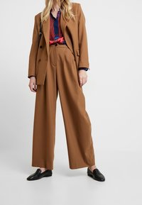 Hnědá saková bunda na míru přes košili s námořnickými a červenými vzory, doplněná širokými hnědými kalhotami a černými plochými botami.