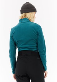 Femme aux cheveux blonds portant un bonnet gris, une veste en polaire turquoise et un pantalon noir, de dos devant un fond blanc.