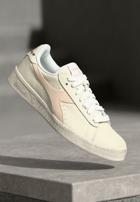 Beige leather sneaker with a soft peach accent, white laces, textured rubber sole, and logo detail on the heel. Rounded toe design.