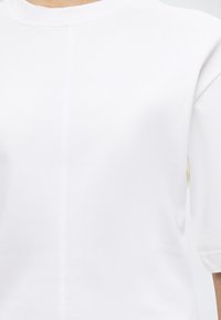 Γυναίκα που φοράει απλό λευκό κοντομάνικο μπλουζάκι με στρογγυλή λαιμόκοψη και κάθετη ραφή κατά μήκος του κέντρου μπροστά.