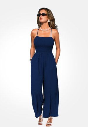 Donna che indossa una tuta senza maniche blu navy a gamba larga, occhiali da sole, collana di conchiglie, orecchini a forma di fiore e tacchi aperti, in piedi con le mani nelle tasche.