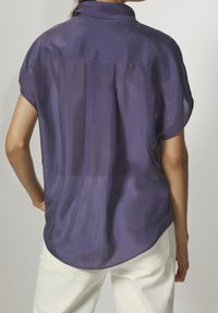 Donna che indossa una blusa trasparente viola a maniche corte con colletto e pantaloni bianchi, vista di spalle su uno sfondo uniforme.
