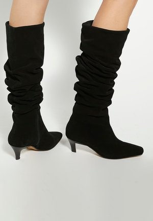 Black slouchy knee-high boots made of suede, featuring a pointed toe and a low kitten heel. Side zipper for easy wear.