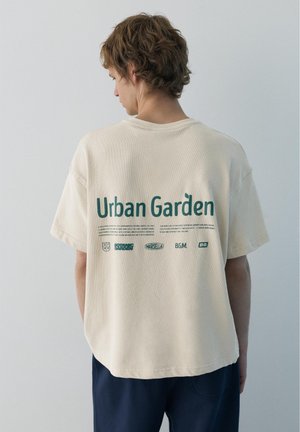 Mladý človek so svetlohnedými kučeravými vlasmi, oblečený v krémové tričko „Urban Garden“ a tmavé nohavice, stojaci tvárou k jednoliatej svetlosivej stene.