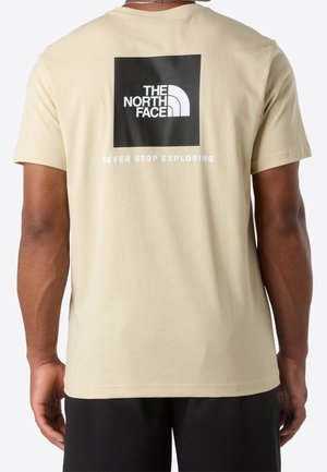 Osoba nosząca beżowy t-shirt z czarnym kwadratowym logo i białym tekstem "THE NORTH FACE" oraz "NEVER STOP EXPLORING" na plecach.
