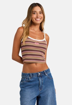 Jeune femme portant un crop top rayé marron, rose et jaune et un jean bleu taille haute, souriant devant un fond uni.