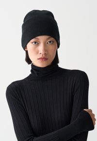 Černá žebrovaná pletená čepice se zaoblenou korunou a decentním detaily značky. V kombinaci s černým žebrovaným rolákem, který je také těsně přiléhavý.