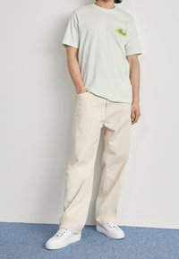 Ljust grön kortärmad t-shirt med en bröstgrafik, i kombination med lösa beige byxor och vita sneakers. Enkelt och avslappnat design.