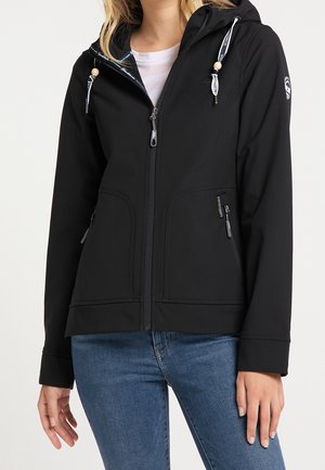 Kvinde iført en sort lynlåset hættejakke med snore, kombineret med blå jeans, stående mod en ensfarvet lys baggrund.