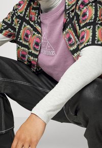 Camisetta nera a maniche corte con grafiche colorate, sovrapposta a una maglietta a girocollo di un leggero colore viola chiaro con logo a triangolo, abbinata a jeans scuri.