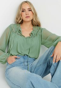 Blusa leggera verde chiaro, trasparente, con scollatura arricciata, texture a pois, maniche lunghe e lacci, abbinata a jeans blu a vita alta.