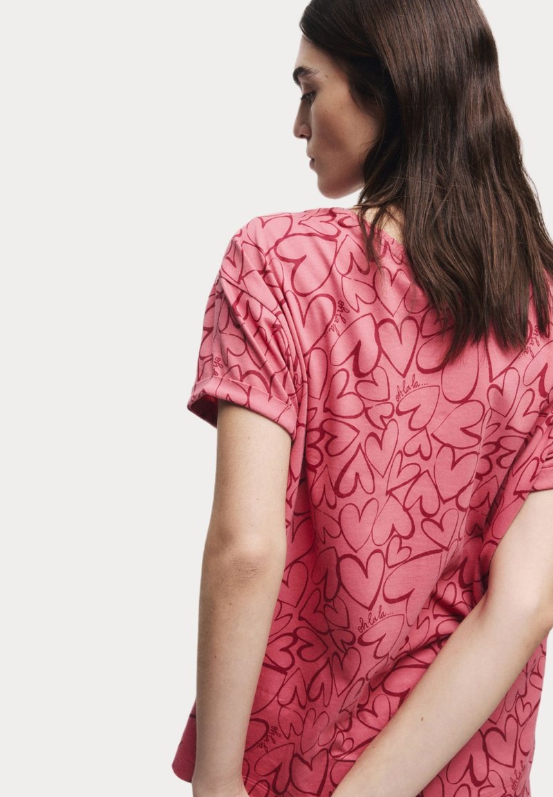Roze T-shirt met korte mouwen, voorzien van een hartjespatroon en subtiele tekstdetails. Zachte stof, ontspannen pasvorm en opgerolde mouwzoom.