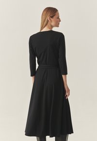 Zwarte jurk met lange mouwen en een taillestrik, voorzien van een geplooemde zoom en een gladde textuur. De achterkant heeft een afgeronde halslijn en een aansluitende pasvorm.
