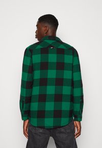 Camicia in flanella a quadri verdi e neri con colletto a punta, polsini abbottonati e un piccolo dettaglio con logo sul retro.