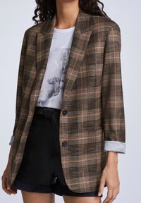 Osoba ubrana w brązową kraciastą marynarkę na białej koszulce z nadrukiem oraz czarne szorty z wysokim stanem, z podwiniętymi rękawami marynarki.