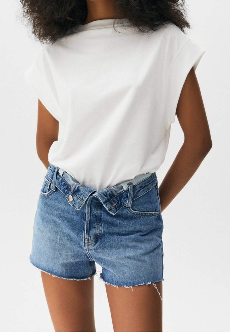 Valkoinen lyhythihainen paita yhdistettynä repeytyneisiin sinisiin farkkuhousuihin. Paidassa on rento leikkaus; shortseissa on napp Closure ja vyölenkit.