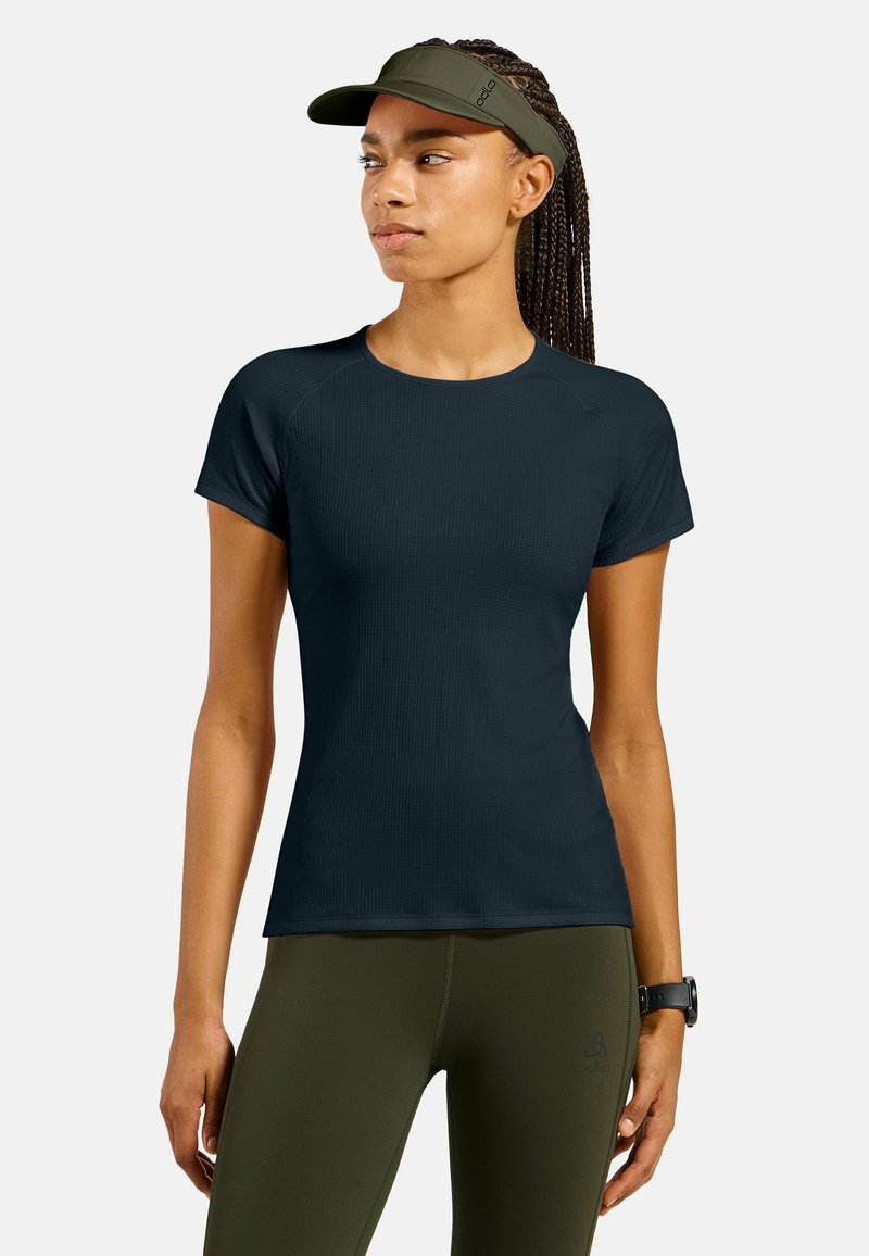 Frau mit geflochtenem Haar, die ein schwarzes, kurzärmliges Sportshirt, olivgrüne Leggings, ein Sportvisier und eine Fitnessuhr trägt und zur Seite blickt.