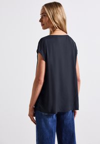 Camicetta nera a maniche corte con orlo arrotondato, texture liscia e vestibilità rilassata, abbinata a jeans blu a vestibilità ampia.
