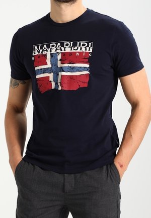 Uomo che indossa una maglietta blu navy con bandiera norvegese consumata e scritta "Napapijri", abbinata a pantaloni grigio scuro, con le mani nelle tasche.