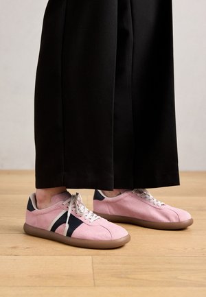 Light pink suede sneakers with navy accents and a cream sole. Rounded toe, white laces, and a low profile design. Worn with black wide-leg pants.