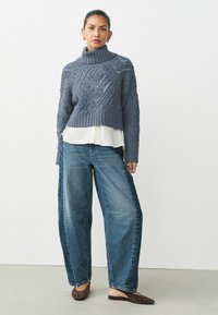 Tjockstickad marinblå tröja med hög krage och mönstrad textur, lagd över en vit skjorta; matchad med vida ljusblå jeans och bruna ballerinaskor.