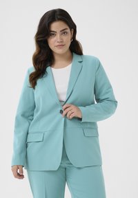 Světle modrý blazer s klopami, zapínání na jeden knoflík a dvěma předními kapsami. Hladká tkanina a přiléhavý střih. Pod ním bílý top.