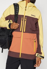 Färgglad parkas med en gul överdel, mörkbrun mittdel och orange nedre del. Har en framdragkedja, två bröstfickor och justerbara ärmslut.