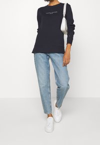 Marineblauwe sweatshirt met wit logo, losse pasvorm, geribde manchetten. Lichtblauwe jeans met een losse snit, gecombineerd met witte sneakers en een zilveren handtas.