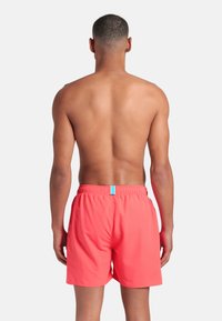 Uomo con capelli corti, a torso nudo, che indossa pantaloni da bagno corallo brillanti, in piedi rivolto di spalle contro uno sfondo bianco semplice.