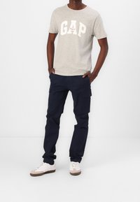 Mand iført en lys grå GAP t-shirt, marineblå cargo-bukser med klaplommer og hvide sneakers med beige detaljer, stående med hænderne i lommerne.