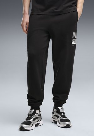 Person iført sorte Puma joggingbukser og sorte og sølvfarvede Puma sneakers, der står mod en grå baggrund.