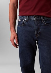 Tumesinised teksapüksid regulaarse istuvusega. Omavad nööpidega lahust, klassikalist viie taskuga disaini ja punast logoga plaastrit vöörihmil.