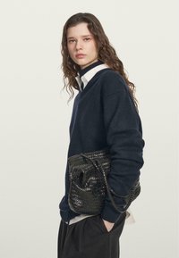 Donkerblauwe trui met een V-hals, gecombineerd met een witte blouse met kraag. Houdt een zwarte gevlochten handtas met een gestructureerde afwerking vast.