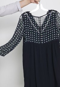 Hand som håller en svart klänning med genomskinliga långa ärmar och prickigt mönster på den övre delen, upphängd på en vit galge.